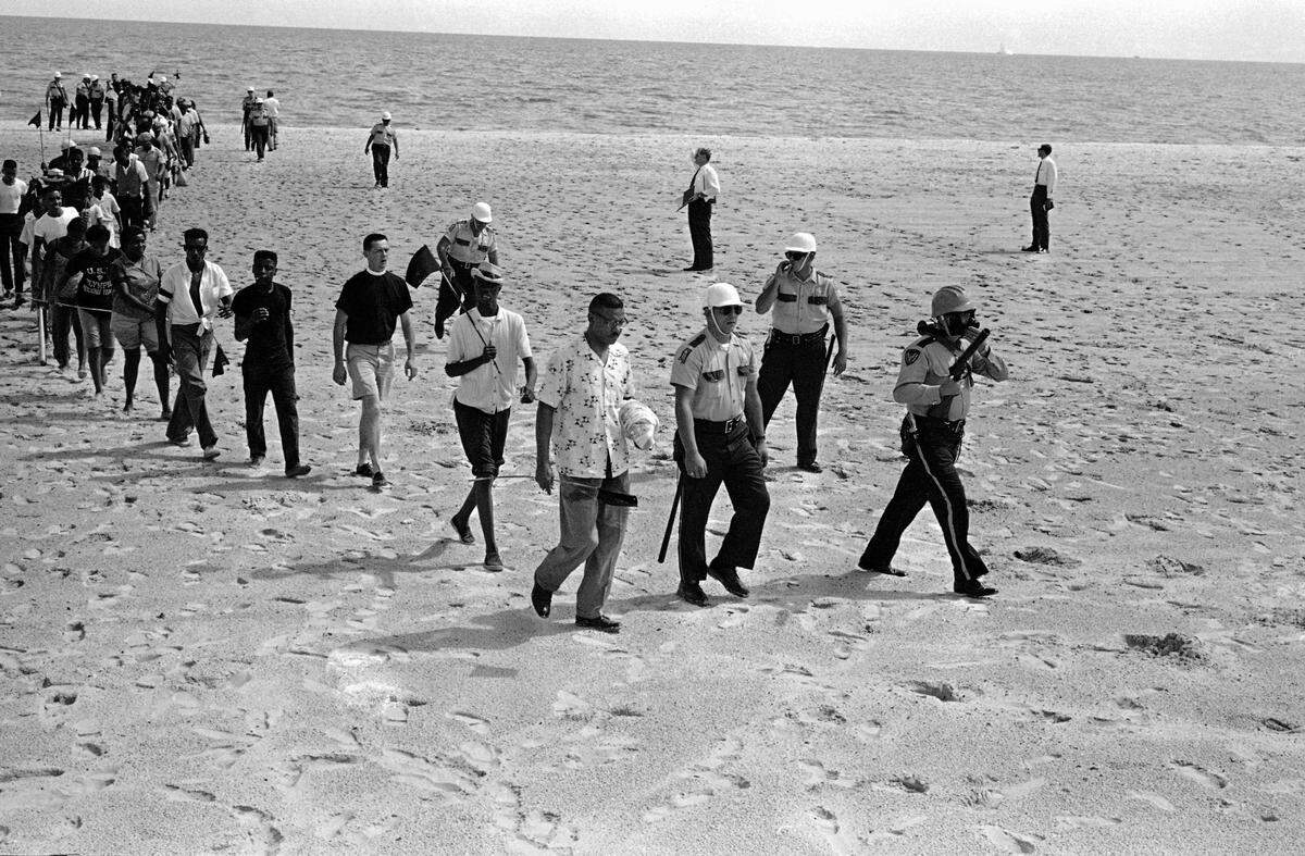 Black and white demonstrators, 65-strong and under arrest, troop off beach at Biloxi, June 23, 1963 after staging unsuccessful attempt to desegregate the Gulf Coast beach. Group was led by Dr. Gilbert Mason (center, foreground) and white minister, Rev. R.G. Gallagher, Biloxi (third in line with shorts). The group was arrested for trespassing on private property.
