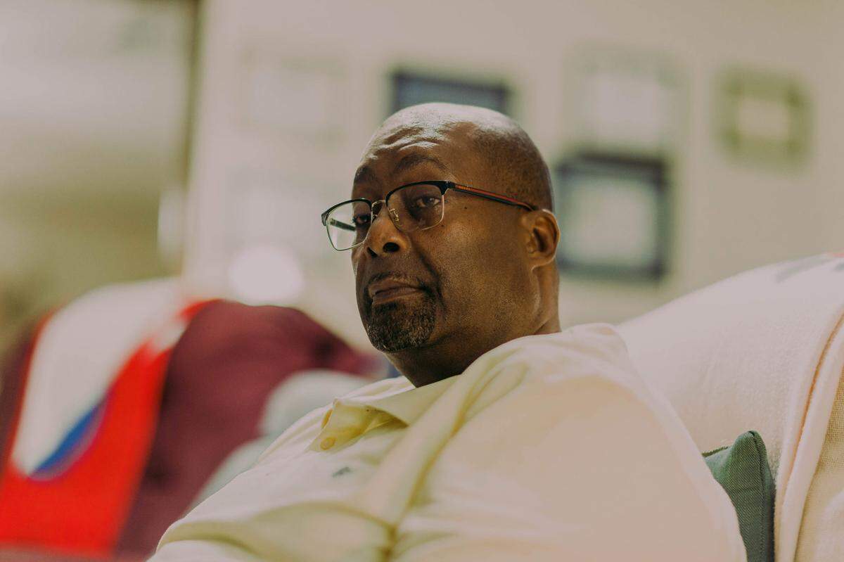 A photo of Gerald Laird, a bald, Black man with a goatee and a cream-colored shirt, sitting on a couch in his living room. Framed certificates are out of focus behind the man.