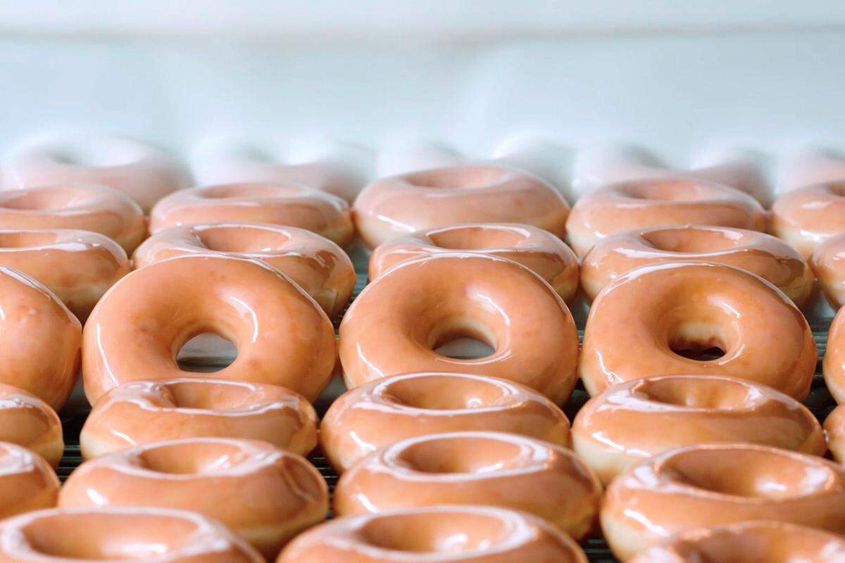 A large batch of freshly made original glazed doughnuts are shown on a wire rack. They are a golden brown color with a shiny glaze coating, and the ones in the back are starting to be coated with a white icing.