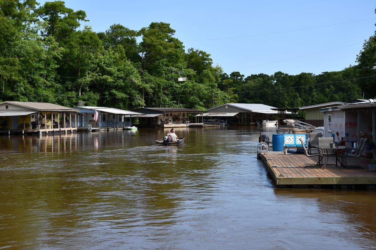 Two people ride in a small motorboat along a river lined with floating houses, porches, and American flags on a sunny day.