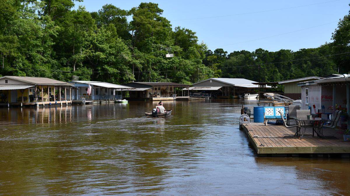 Two people ride in a small motorboat along a river lined with floating houses, porches, and American flags on a sunny day.