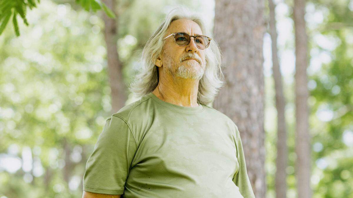 A photo of John Cook, a White man with gray shoulder-length hair, a gray beard, glasses and a green shirt. Cook is looking into the distance while standing outdoors under a tree. 