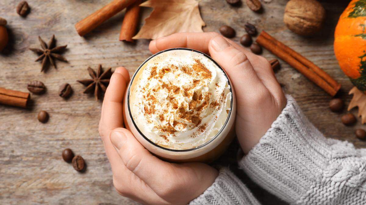 A woman holds a glass of tasty pumpkin spice latte on a wooden table.